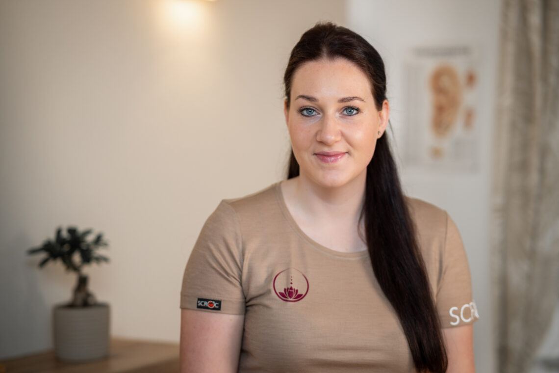 Sarah Schedlberger steht in ihrer Praxis. Sie trägt ihre dunklen langen Haare halb offen. Sie hat ein beiges T-Shirt mit ihrem Praxislogo darauf an.