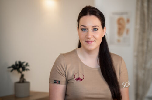 Sarah Schedlberger steht in ihrer Praxis. Sie trägt ihre dunklen langen Haare halb offen. Sie hat ein beiges T-Shirt mit ihrem Praxislogo darauf an.